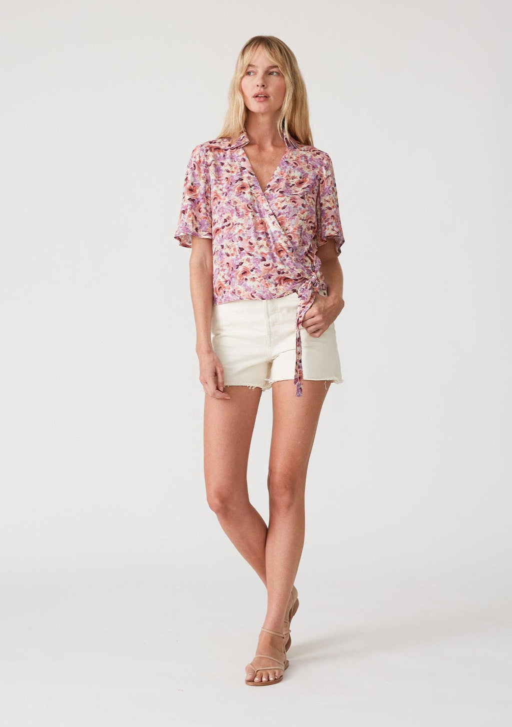 Woman wearing a floral wrap top with side tie and white shorts on a plain background