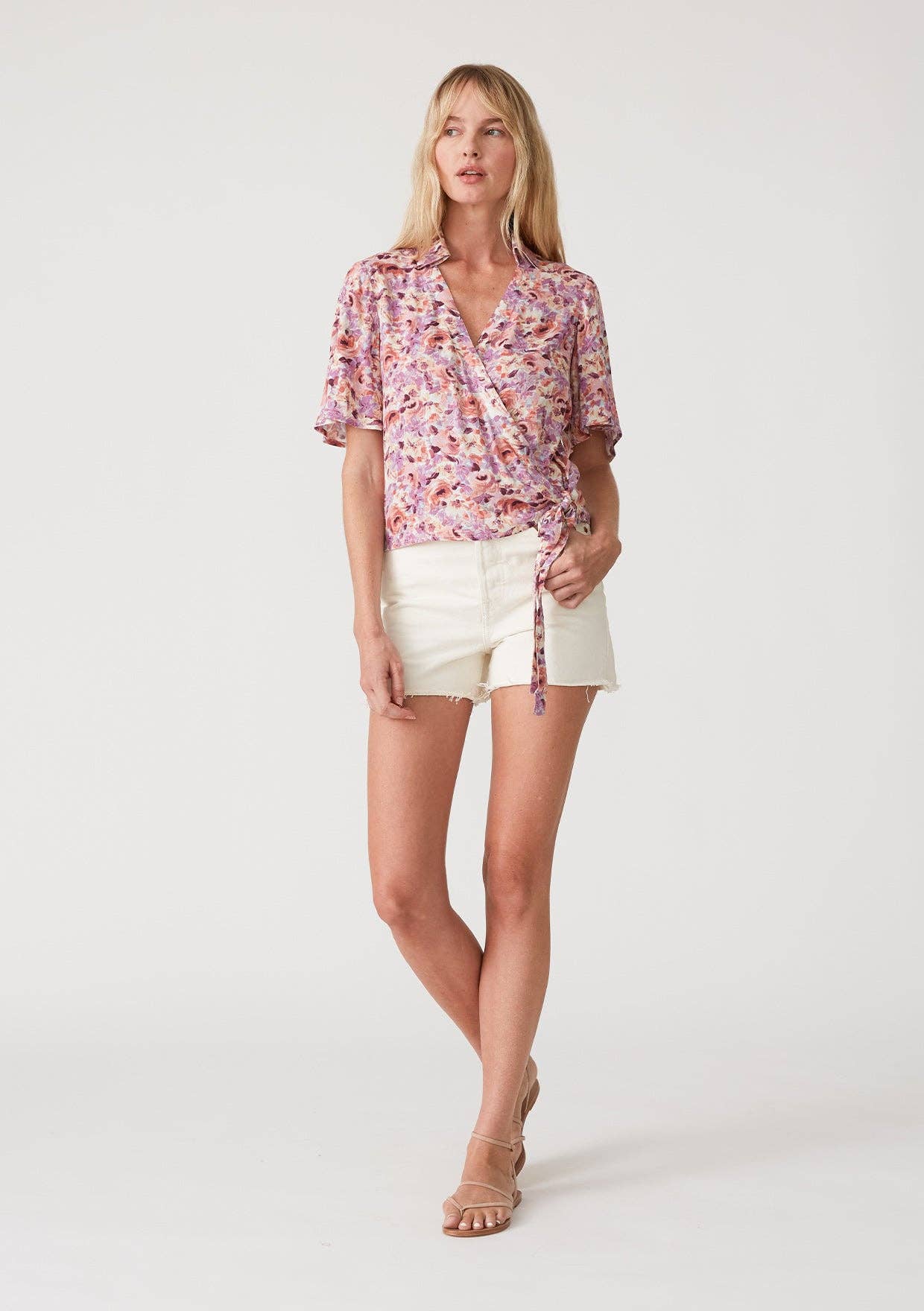 Woman wearing a floral wrap top with side tie and white shorts on a plain background