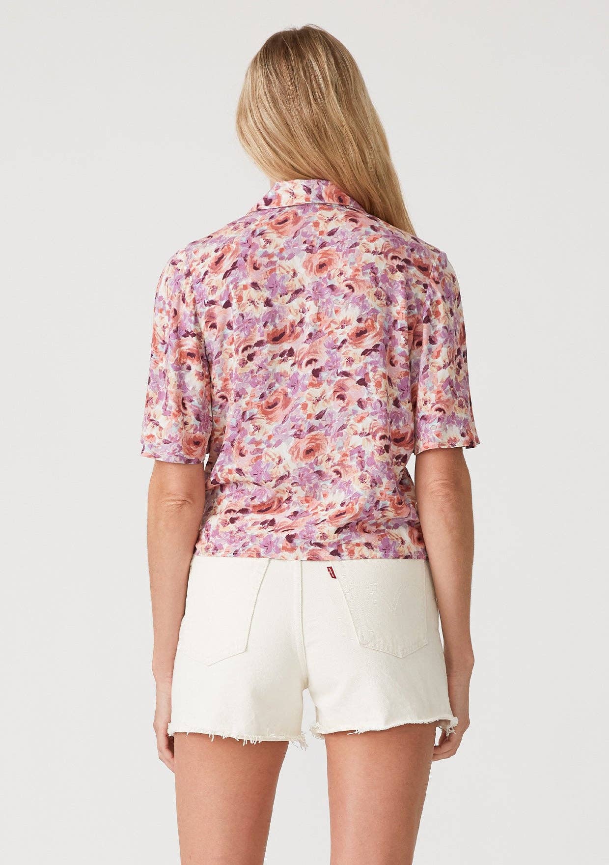 Back view of a woman wearing a floral wrap top with side tie and white shorts on a plain background