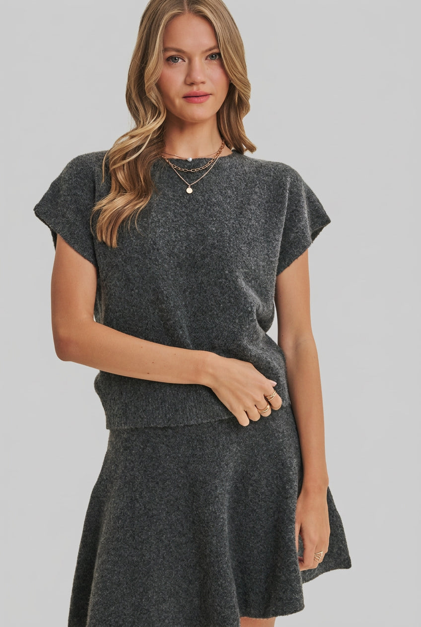 A woman modeling a charcoal knit dolman short-sleeve top and a matching flared skirt against a solid light beige background.