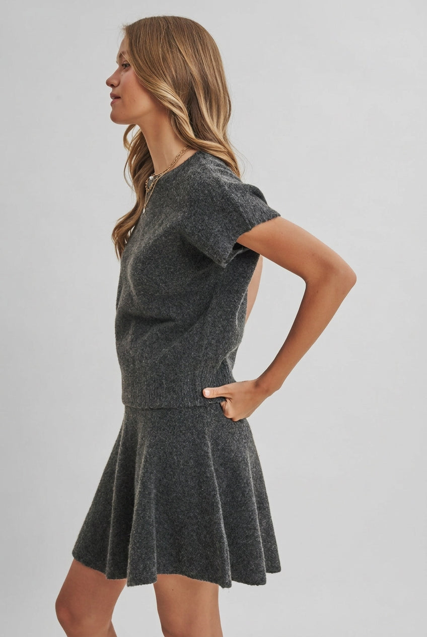 A woman modeling a charcoal knit dolman short-sleeve top and a matching flared skirt against a solid light beige background.