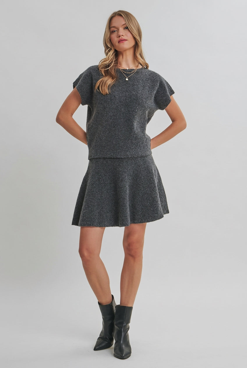 A woman modeling a charcoal knit dolman short-sleeve top and a matching flared skirt against a solid light beige background.