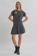 A woman modeling a charcoal knit dolman short-sleeve top and a matching flared skirt against a solid light beige background.