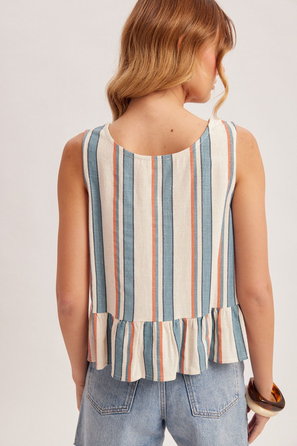 Back view of a woman wearing a striped blue, red and cream tank top with jeans on a plain background