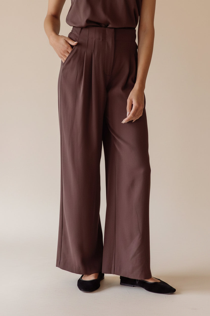 Woman wearing brown wide leg pants standing against a neutral wall.