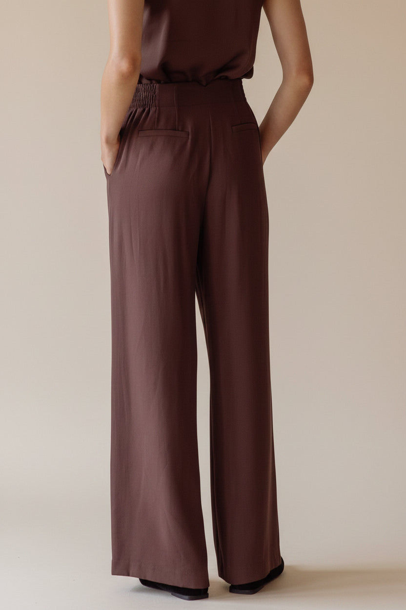 Back view of a woman wearing a brown wide leg pants standing against a neutral wall.