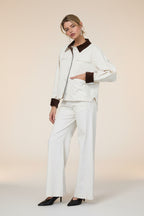 Woman in a white barn jacket with brown accents and white pants standing against a grey wall