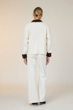 Back view of a woman in a white barn jacket with brown accents and white pants standing against a grey wall