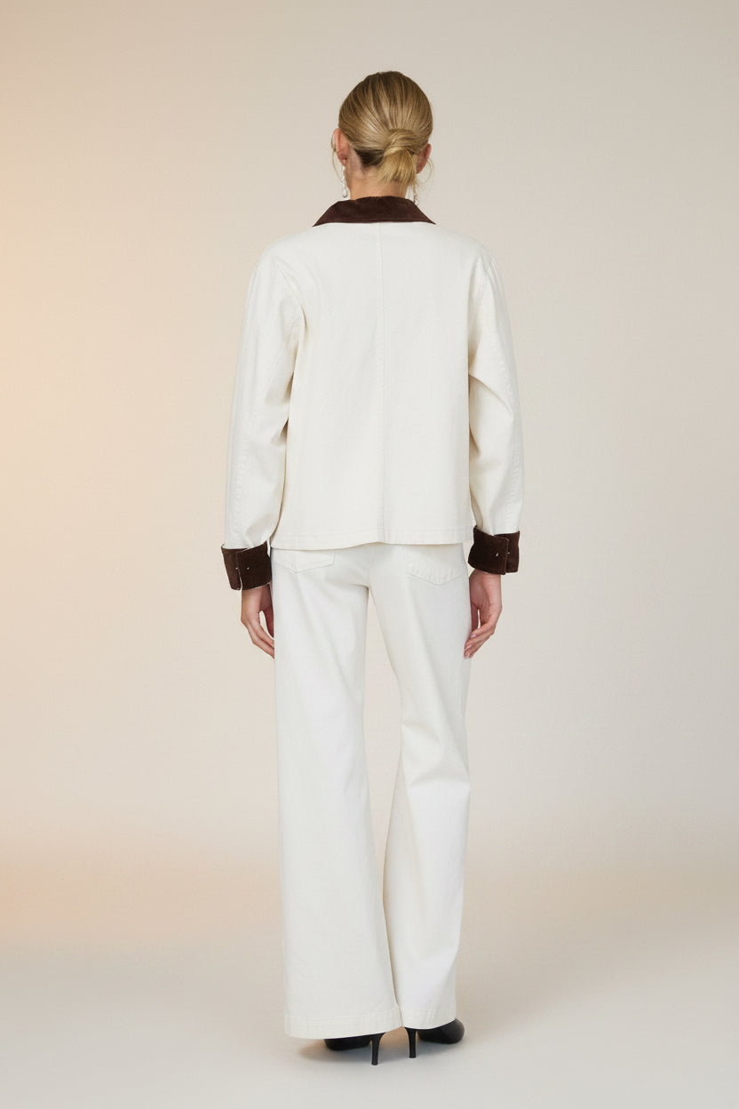 Back view of a woman in a white barn jacket with brown accents and white pants standing against a grey wall