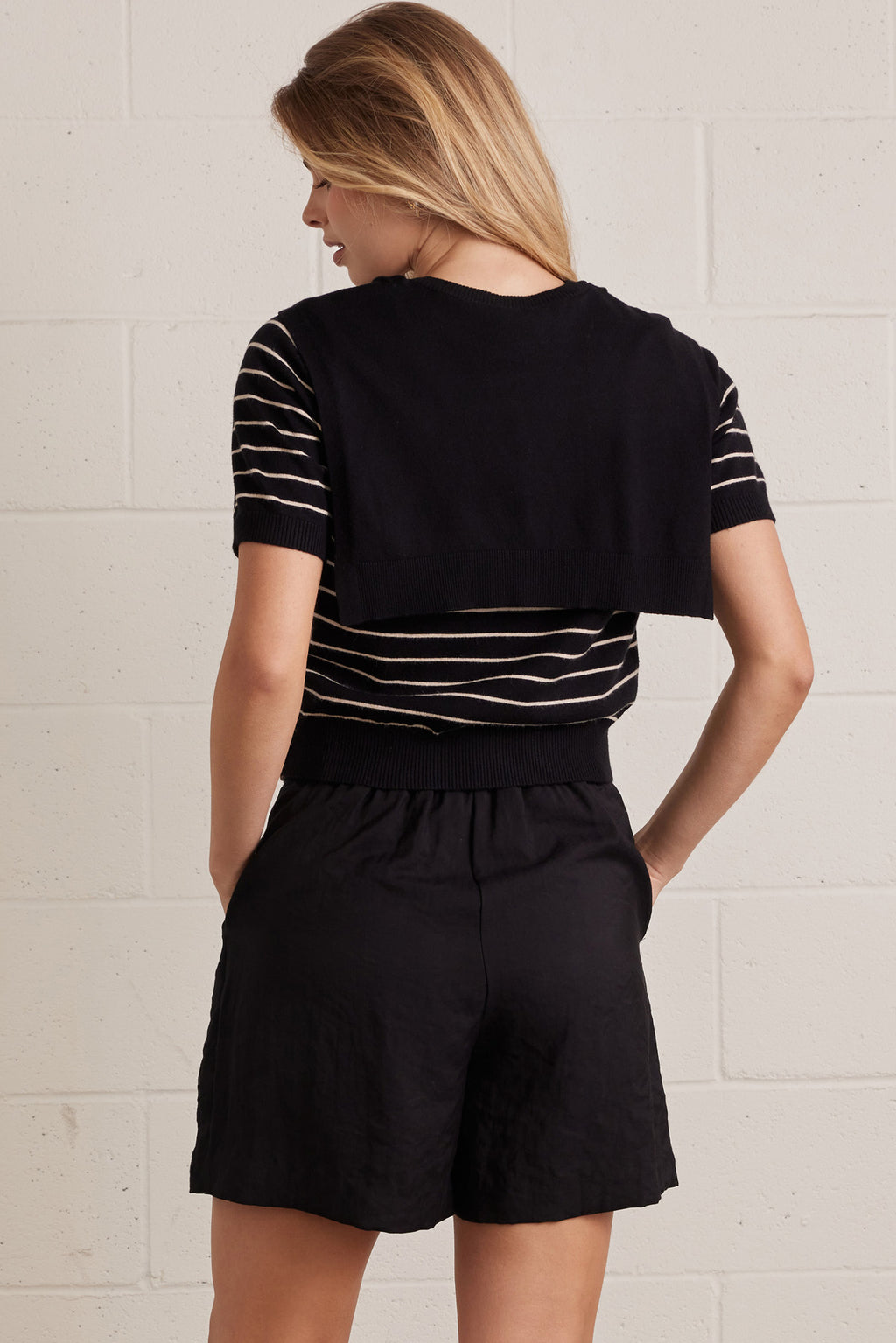 Back view of a a woman wearing a striped short sleeve knit top with an attached layered shoulder shirt style against a grey background
