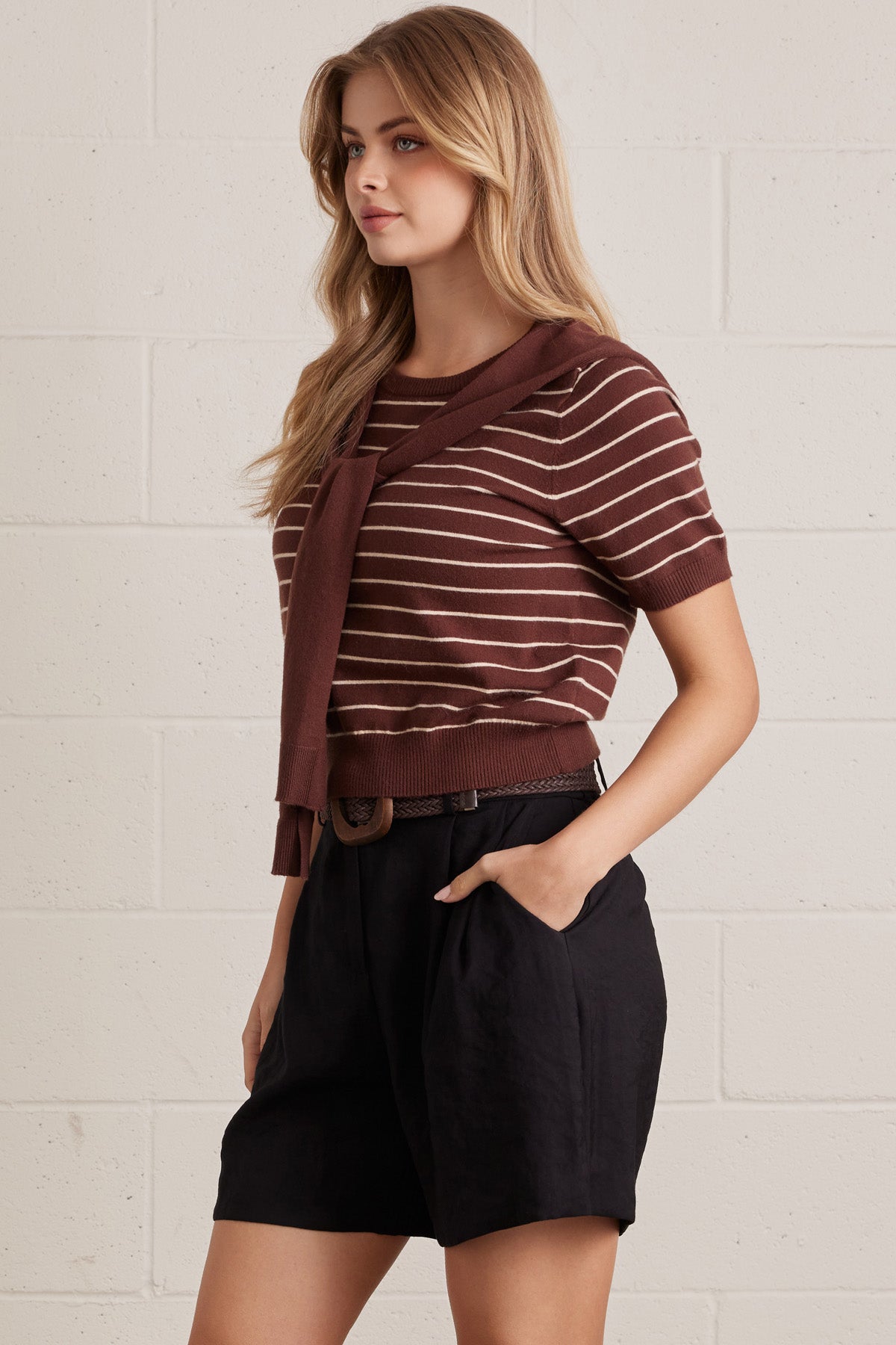 Woman wearing a brown striped shirt with brown layered shirt style scarf and black shorts against a light brick wall.