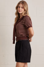 Woman wearing a brown striped shirt with brown layered shirt style scarf and black shorts against a light brick wall.