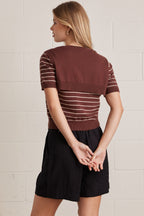 Back view of a woman wearing a brown striped shirt with brown layered shirt style scarf and black shorts against a light brick wall.