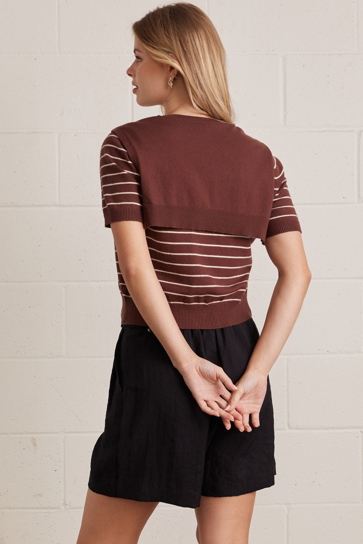 Back view of a woman wearing a brown striped shirt with brown layered shirt style scarf and black shorts against a light brick wall.