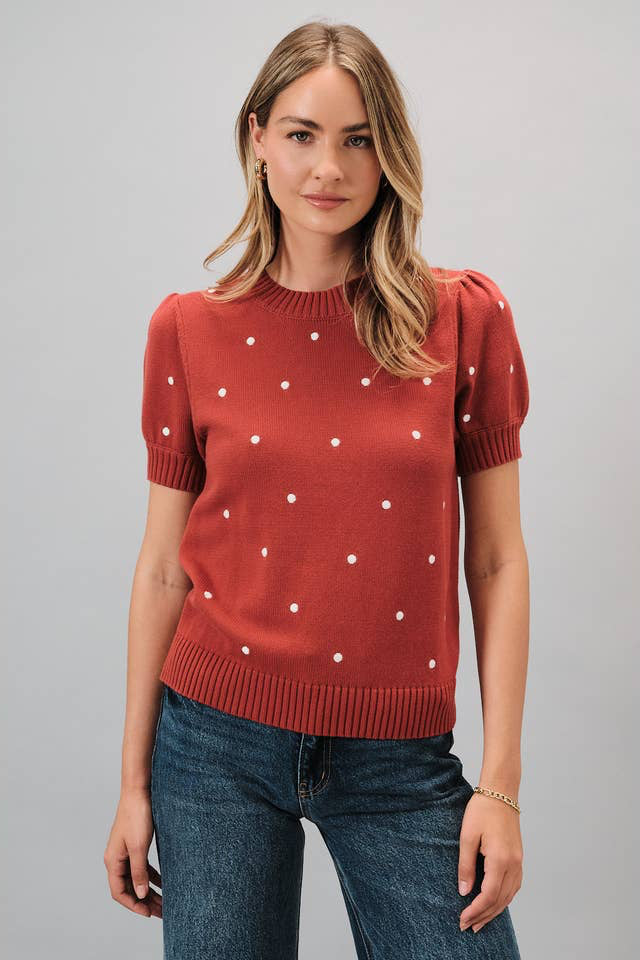 A woman wearing a rust short sleeve knit sweater with ivory polka dots and blue jeans, standing against a solid gray studio background.