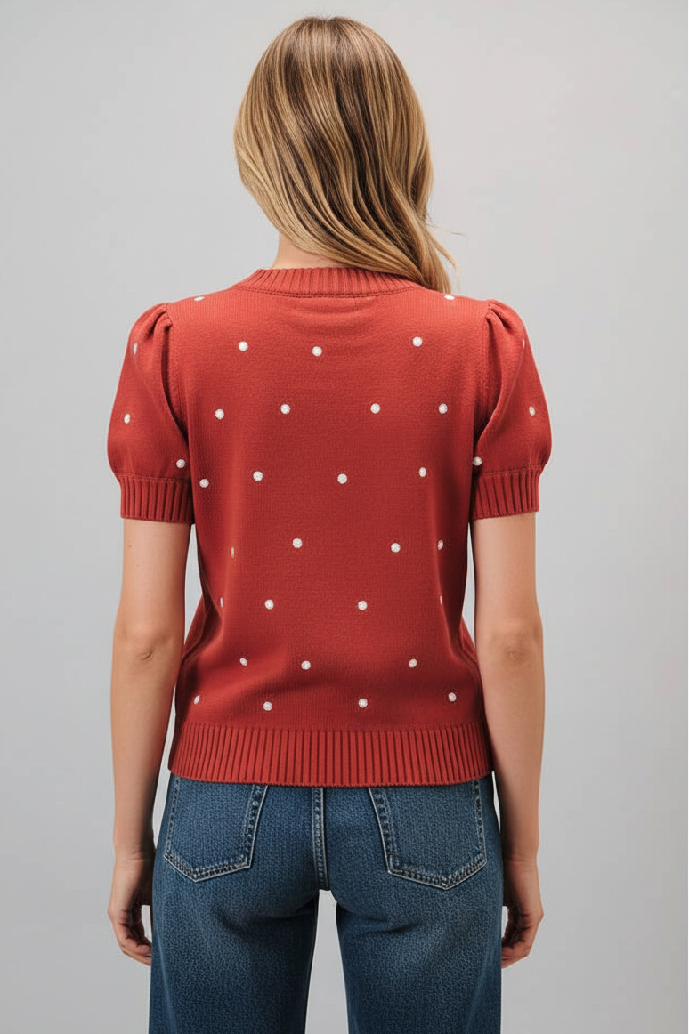 Back view of a woman wearing a rust short sleeve knit sweater with ivory polka dots and blue jeans, standing against a solid gray studio background.