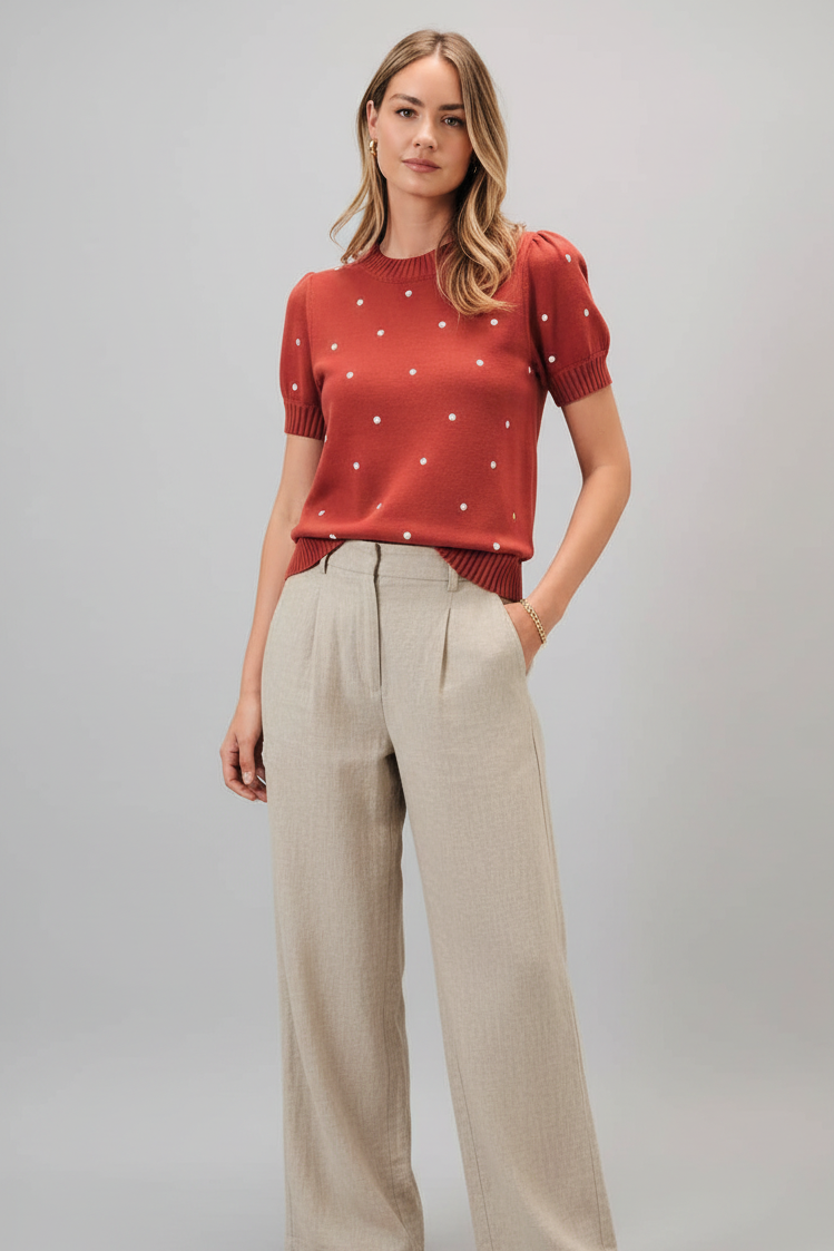 A woman wearing a rust short sleeve knit sweater with ivory polka dots and light linen trouser, standing against a solid gray studio background.