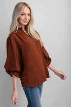 A woman modeling a mocha airflow textured blouse with three-quarter ruffled cuff sleeves and a split neckline, paired with blue jeans against a solid gray background.