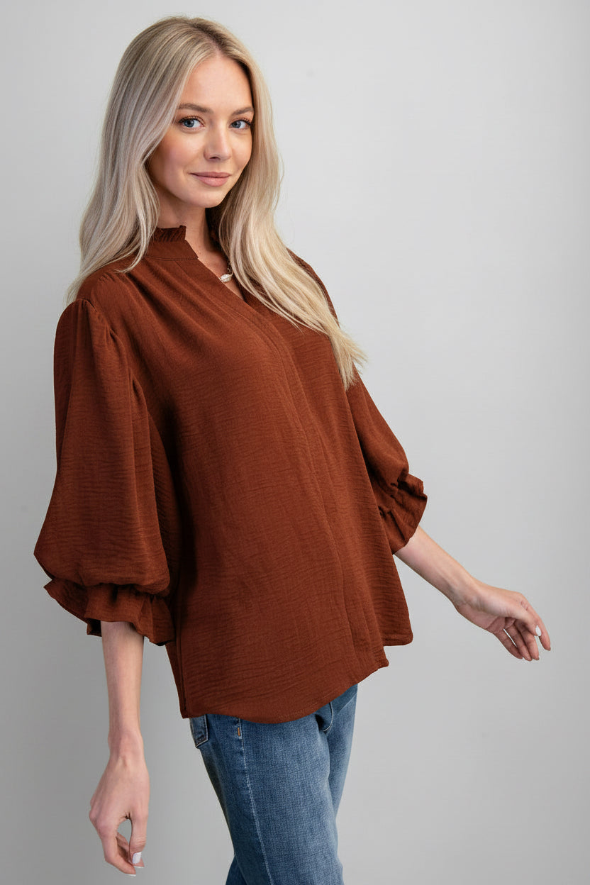 A woman modeling a mocha airflow textured blouse with three-quarter ruffled cuff sleeves and a split neckline, paired with blue jeans against a solid gray background.