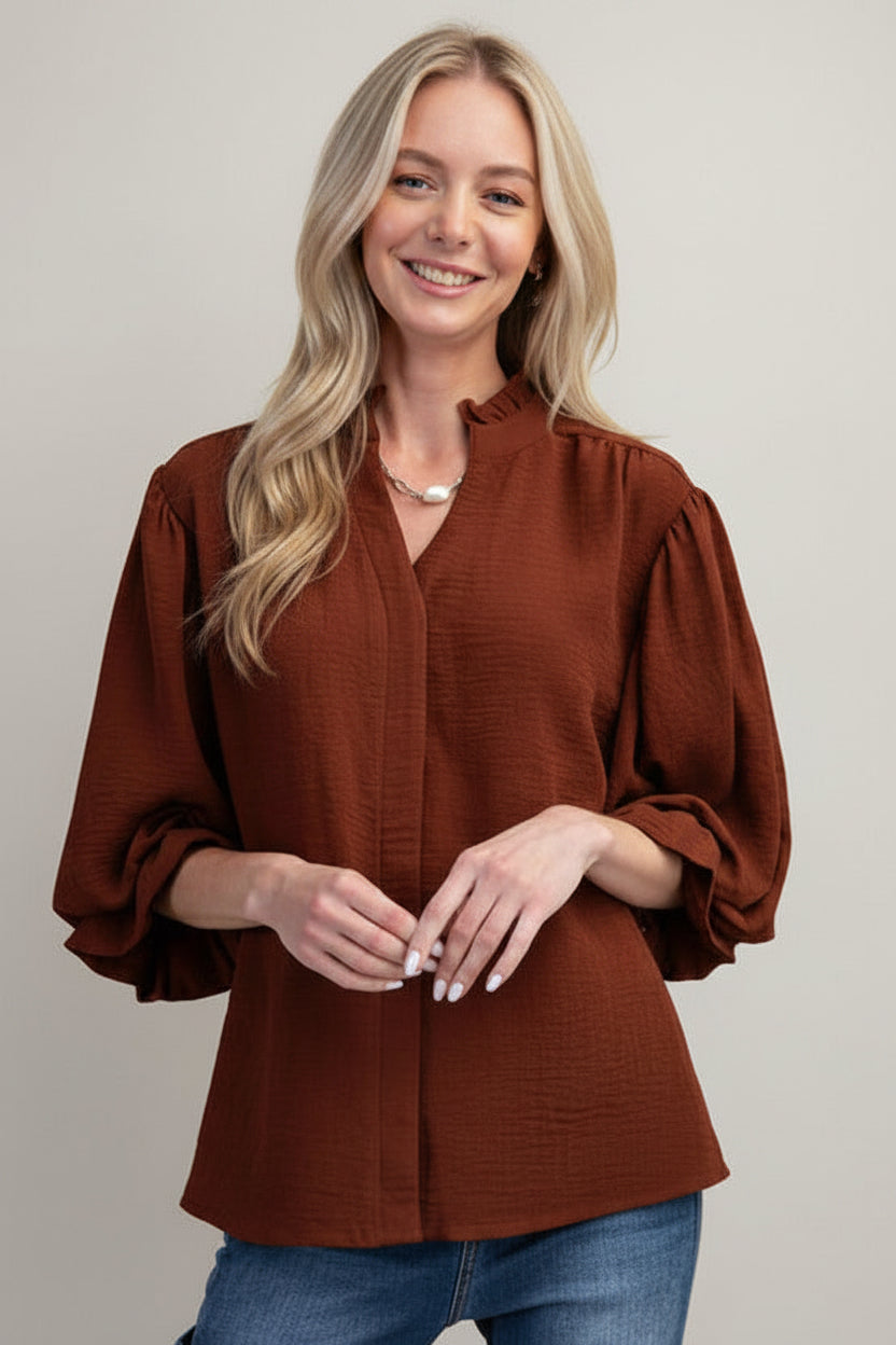 A woman modeling a mocha airflow textured blouse with three-quarter ruffled cuff sleeves and a split neckline, paired with blue jeans against a solid gray background.