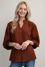 A woman modeling a mocha airflow textured blouse with three-quarter ruffled cuff sleeves and a split neckline, paired with blue jeans against a solid gray background.