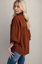 Back view of a woman modeling a mocha airflow textured blouse with three-quarter ruffled cuff sleeves and a split neckline, paired with blue jeans against a solid gray background.