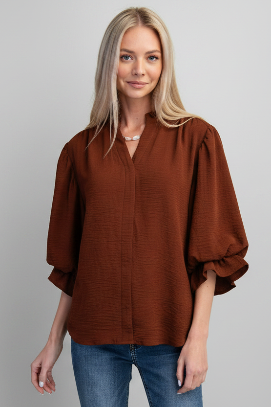 A woman modeling a mocha airflow textured blouse with three-quarter ruffled cuff sleeves and a split neckline, paired with blue jeans against a solid gray background.