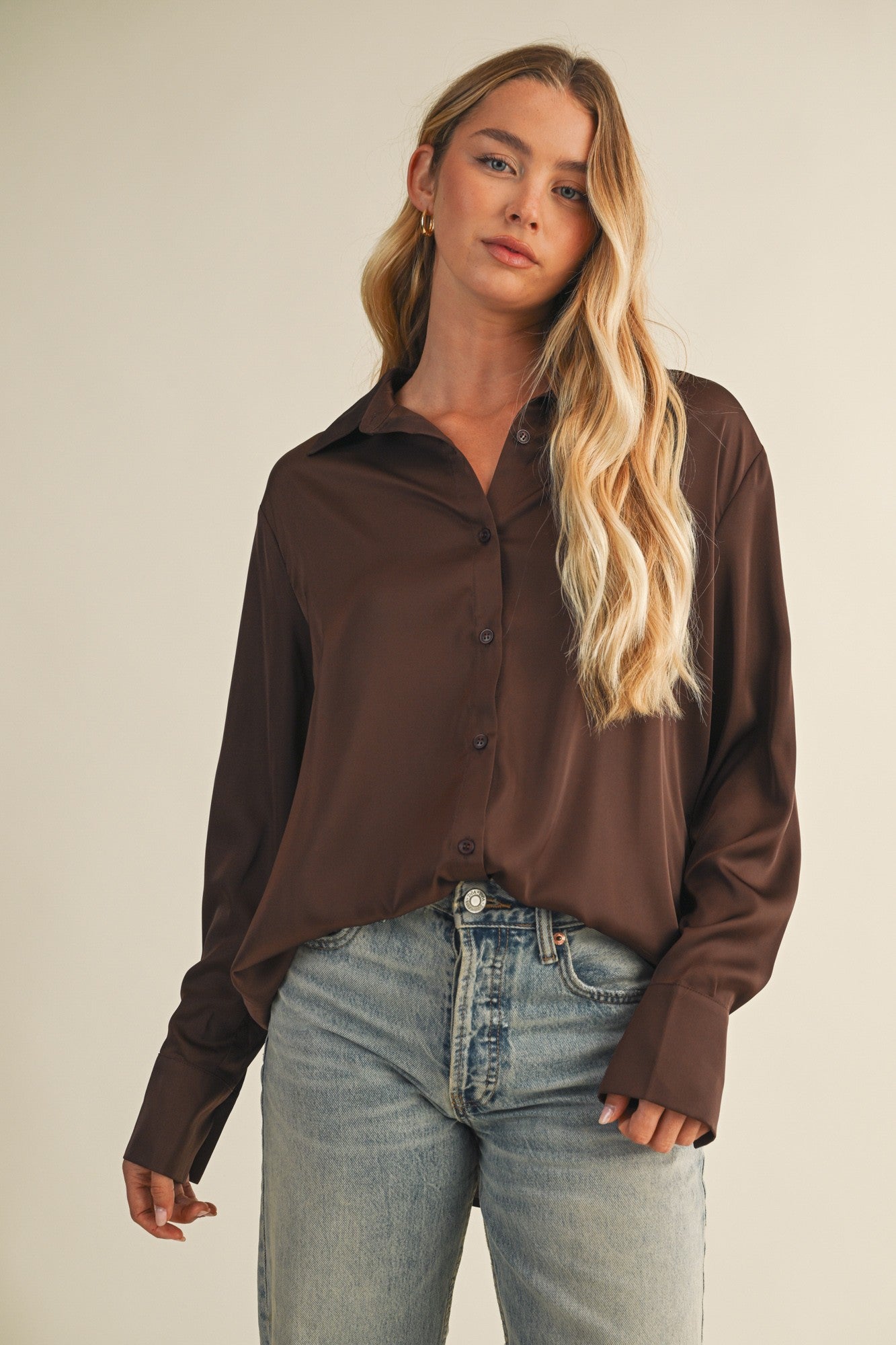Woman wearing a chocolate button-up blouse and blue jeans against a beige background