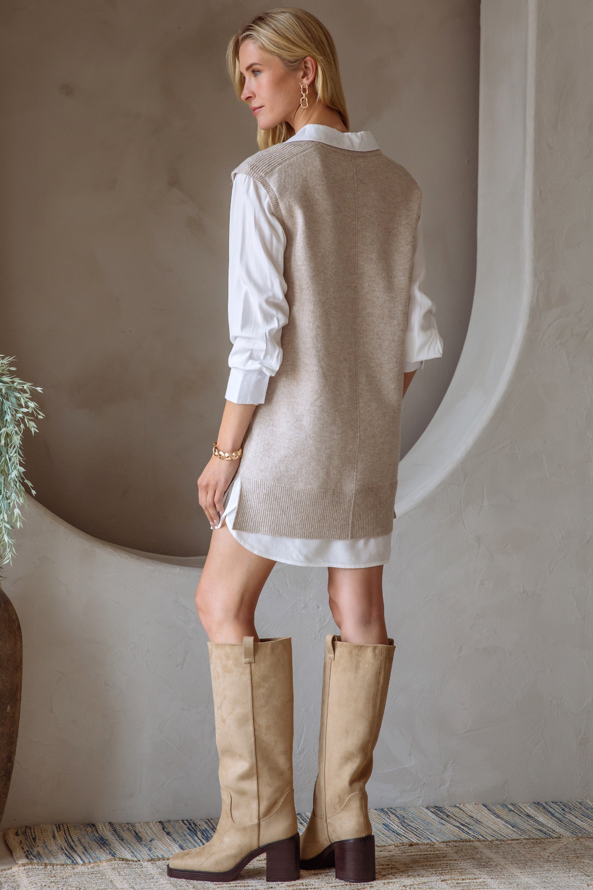 Back view of a woman in a beige and cream layered shirt dress and boots standing next to a neutral wall