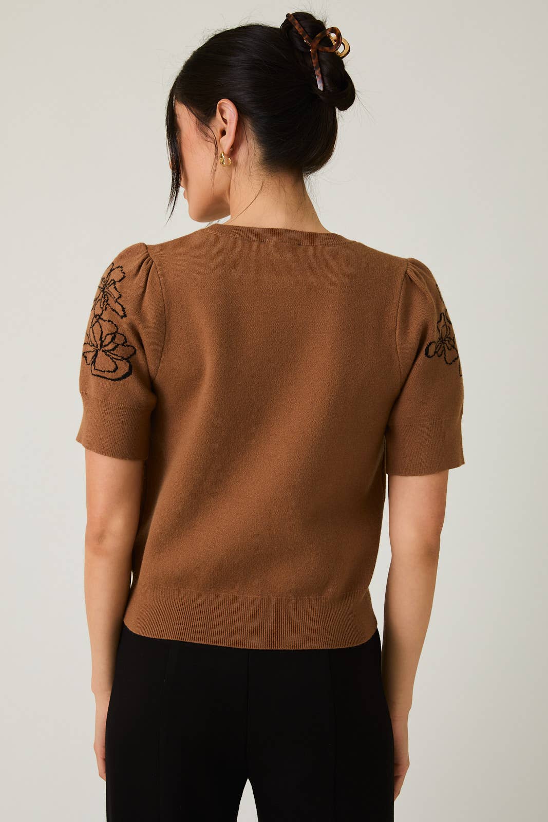 Woman wearing a brown sweater with black flower design on sleeves against a plain background