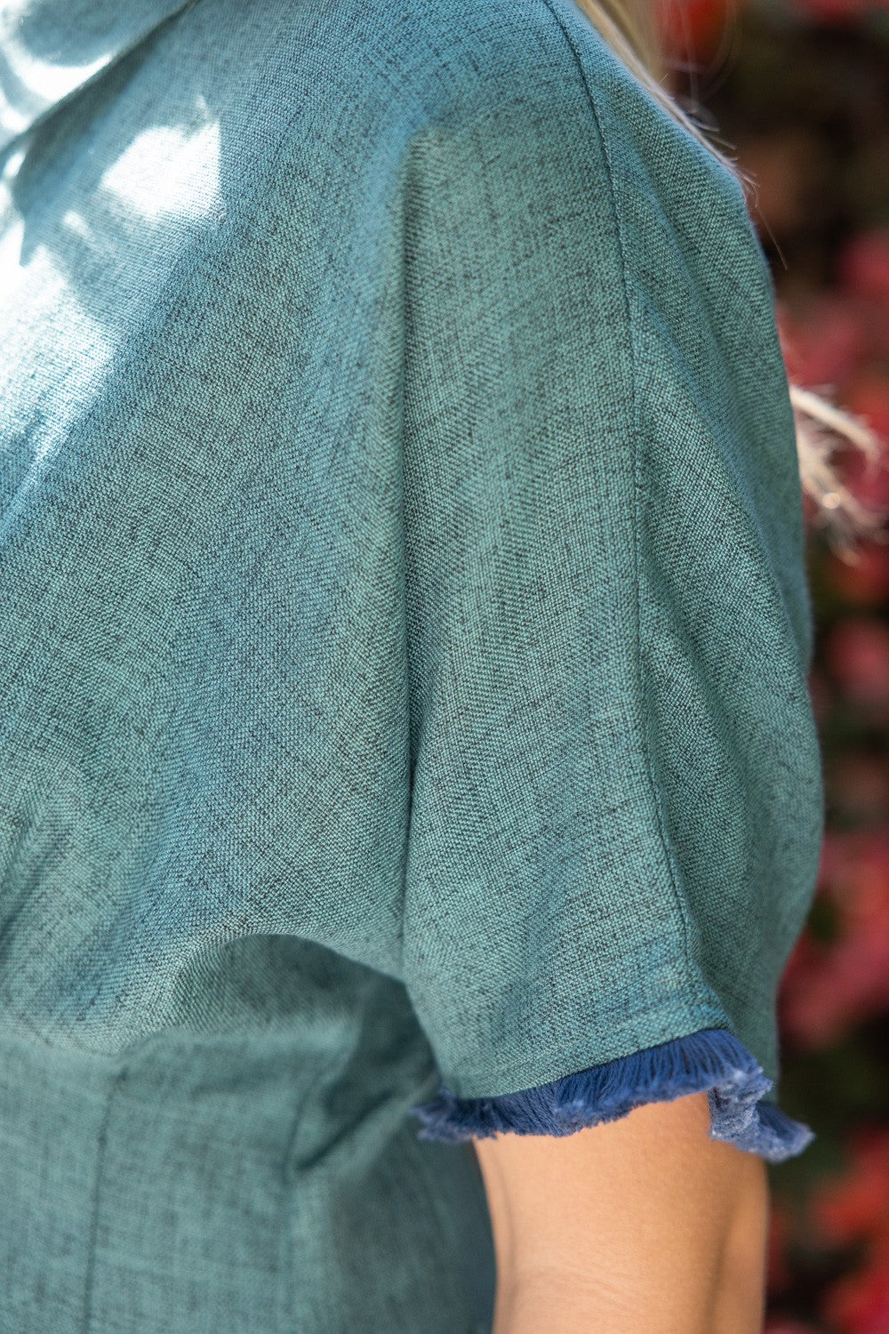 Closeup of a woman wearing a short-sleeve collared button-up shirt in a hunter green color, standing outdoors in front of a background of red and pink flowers.