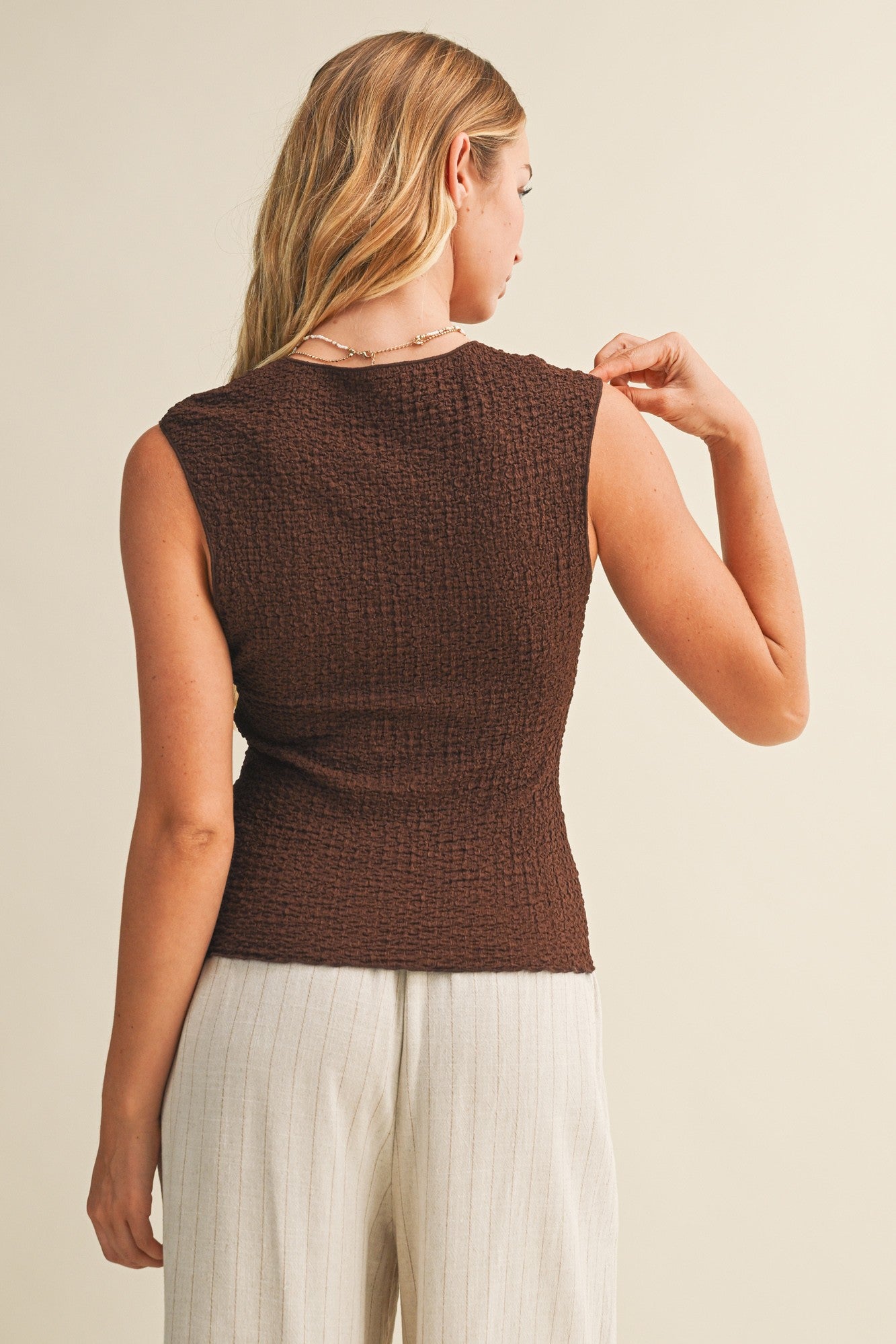 Back view of a woman wearing a brown bubble texture sleeveless top and white pants against a beige background