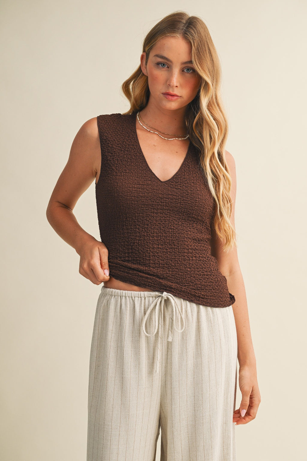Woman wearing a brown bubble texture sleeveless top and white pants against a beige background