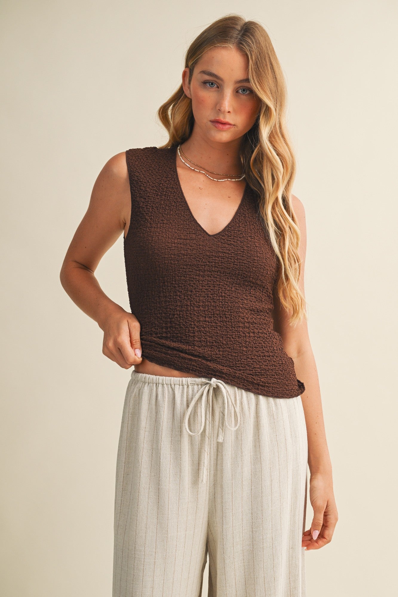 Woman wearing a brown bubble texture sleeveless top and white pants against a beige background