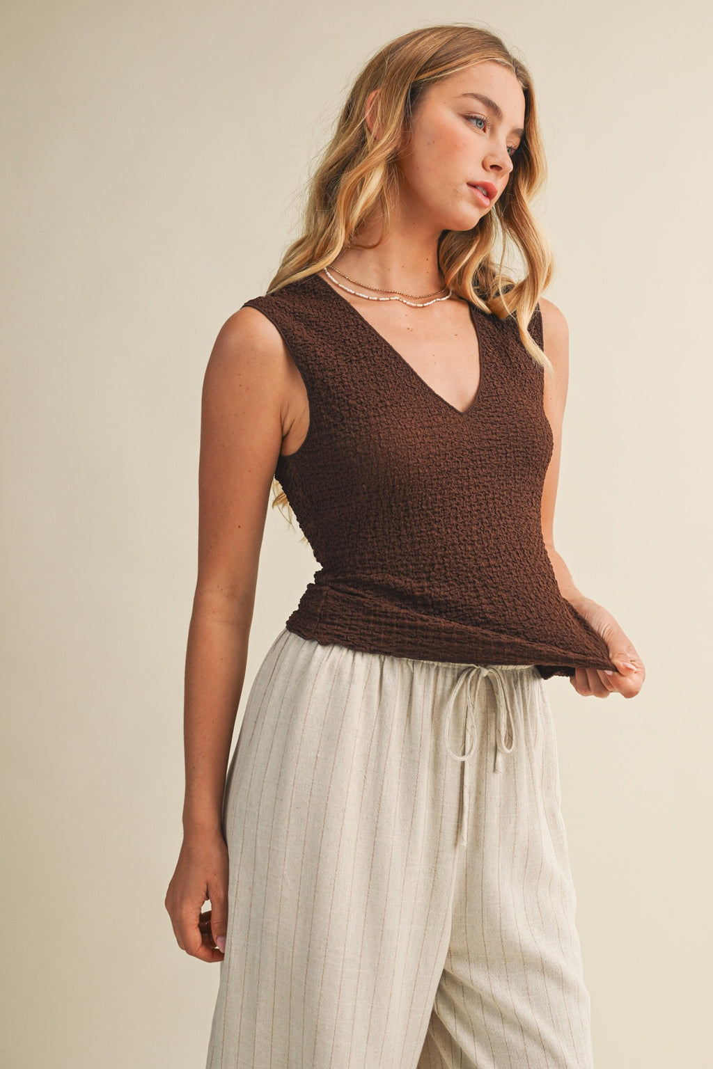 Woman wearing a brown bubble texture sleeveless top and white pants against a beige background