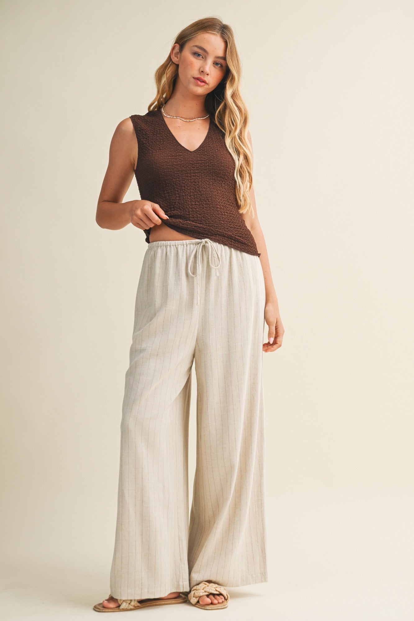 Woman wearing a brown bubble texture sleeveless top and white pants against a beige background