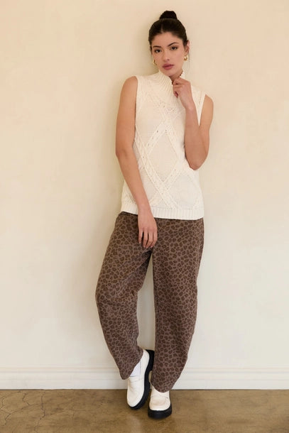 Woman wearing a cream mock neck knitted sleeveless top with brown pants against a beige background