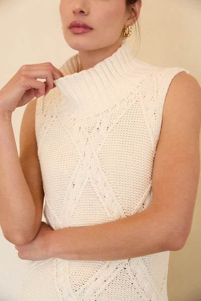Woman wearing a cream mock neck knitted sleeveless top against a beige background