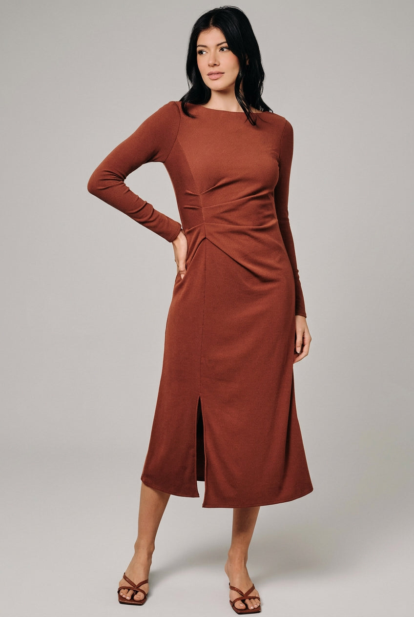 A woman modeling a rust-brown long-sleeve midi dress with a gathered waist and front slit, standing against a neutral beige studio backdrop.
