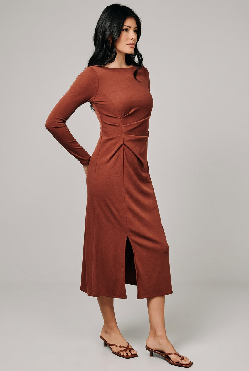 A woman modeling a rust-brown long-sleeve midi dress with a gathered waist and front slit, standing against a neutral beige studio backdrop.
