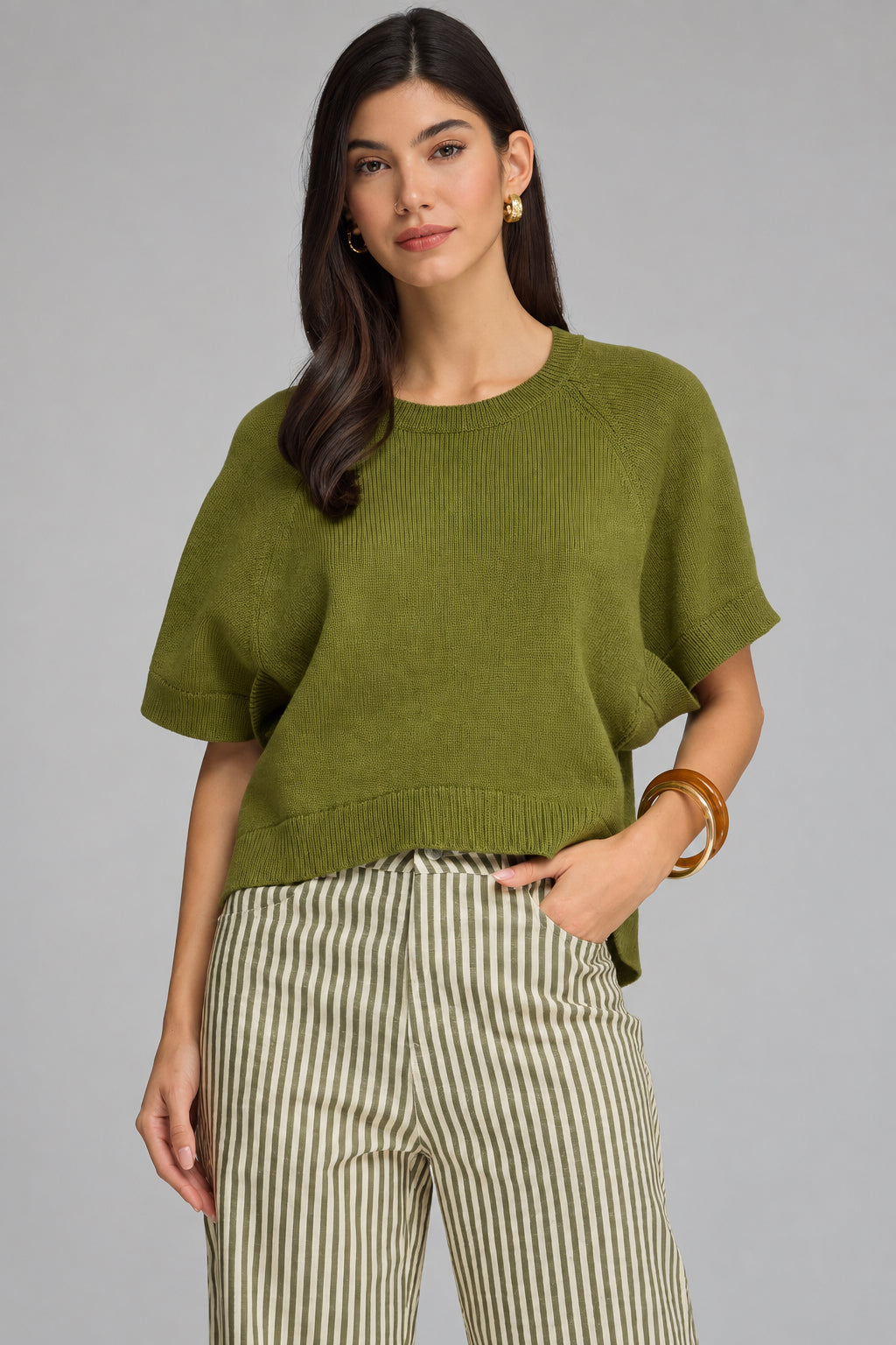 A woman modeling a short-sleeve knit top in olive green, paired with beige and green vertically striped trousers, against a solid gray studio background.