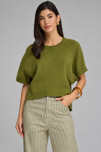 A woman modeling a short-sleeve knit top in olive green, paired with beige and green vertically striped trousers, against a solid gray studio background.