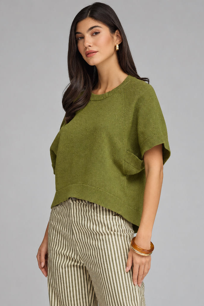 A woman modeling a short-sleeve knit top in olive green, paired with beige and green vertically striped trousers, against a solid gray studio background.