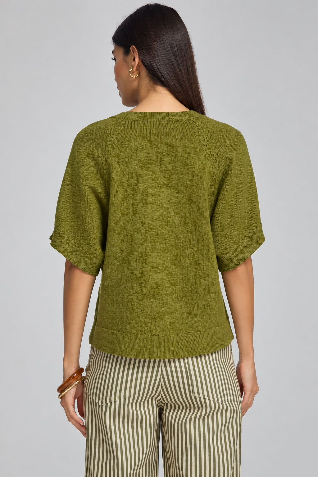 A woman shown from the back wearing a short-sleeve knit top in olive green and striped pants, photographed against a plain gray studio background.