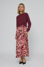 A woman stands indoors wearing a long-sleeve knit sweater in burgundy paired with maxi dress featuring a burgundy and cream floral pattern. 