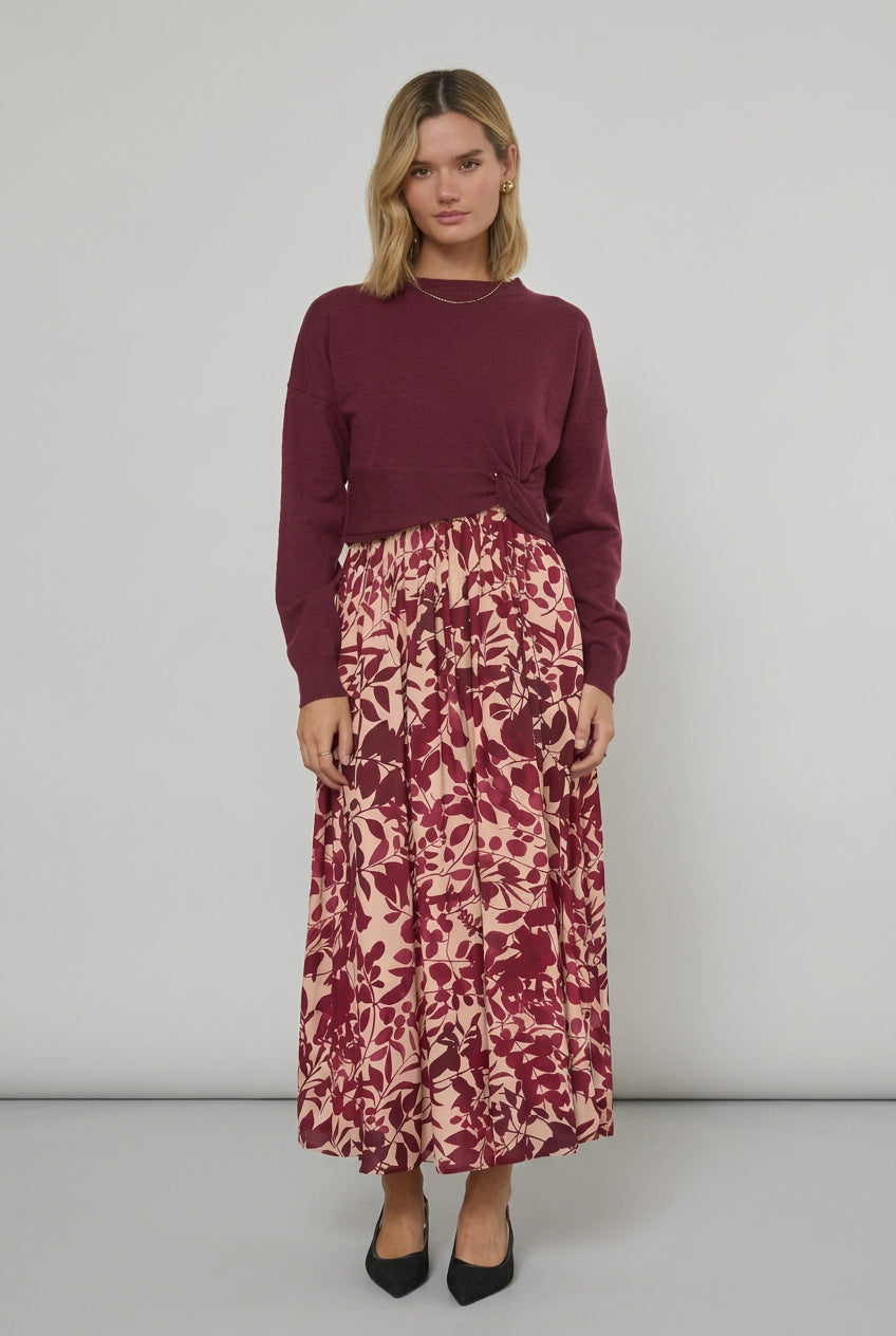 A woman stands indoors wearing a long-sleeve knit sweater in burgundy paired with maxi dress featuring a burgundy and cream floral pattern. She is wearing black pointed-toe heels in a bright room with white curtains.