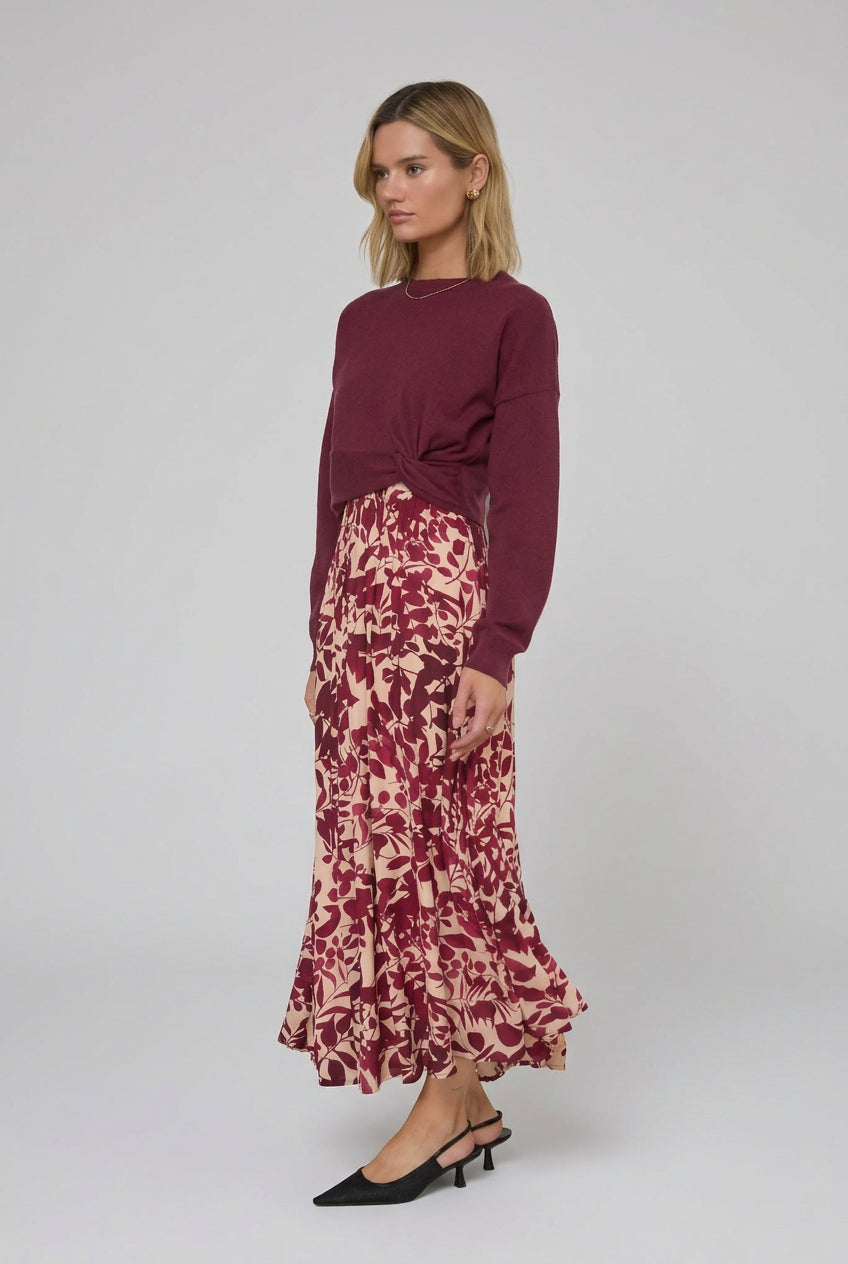 A woman stands indoors wearing a long-sleeve knit sweater in burgundy paired with maxi dress featuring a burgundy and cream floral pattern. She is wearing black pointed-toe heels in a bright room with white curtains.