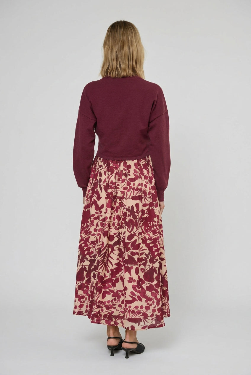  Back view of a woman stands indoors wearing a long-sleeve knit sweater in burgundy paired with maxi dress featuring a burgundy and cream floral pattern. She is wearing black pointed-toe heels in a bright room with white curtains.