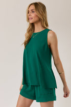 Woman wearing a hunter green A-line  vertical seam tank top and shorts against a plain background