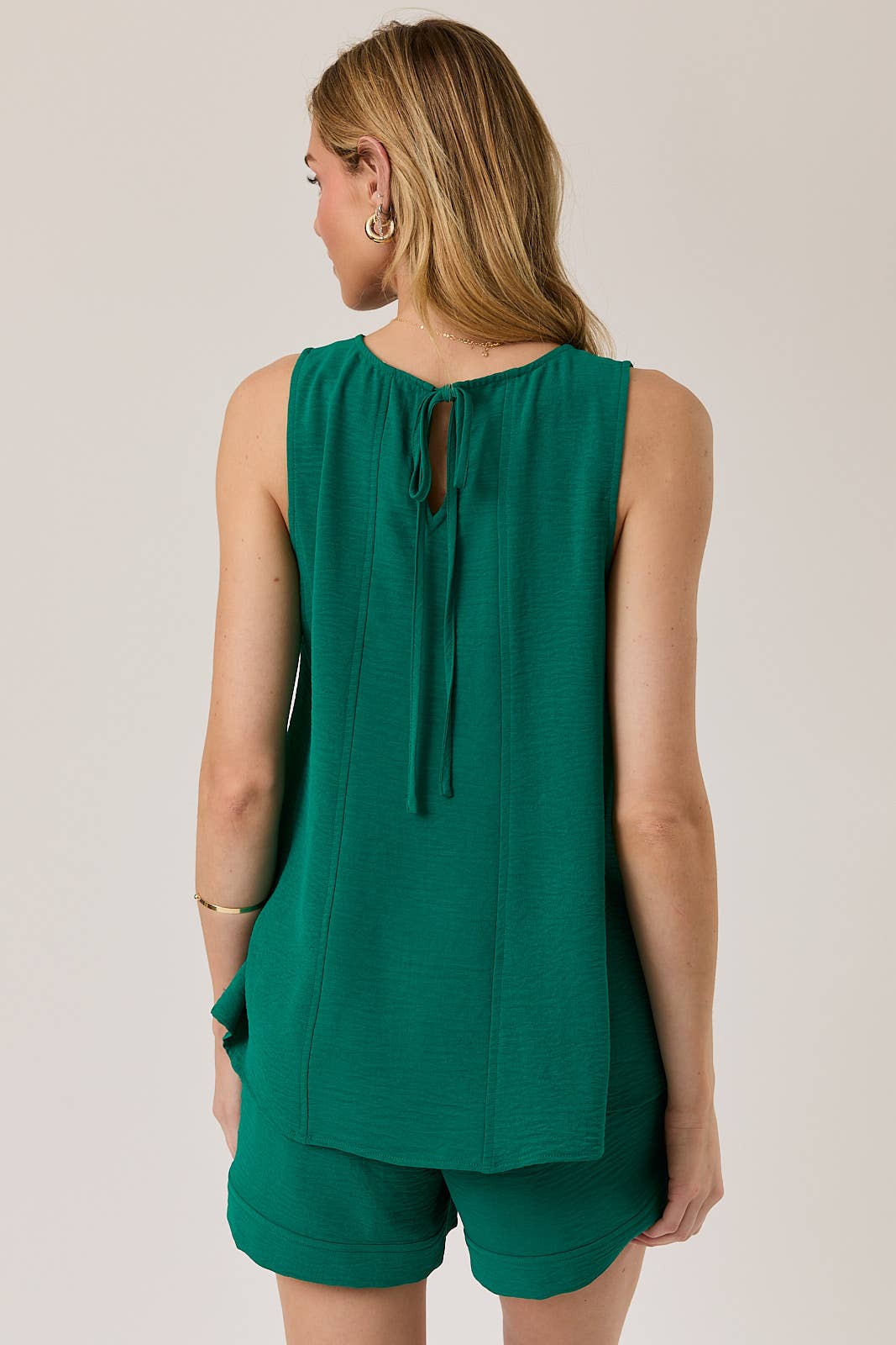 Back view of a woman wearing a hunter green tank top with tie back and shorts against a plain background